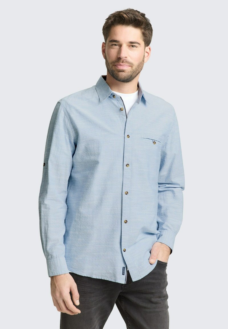 Mand med skæg iført lyseblå skjorte over hvid t-shirt og mørke bukser, stående med den ene hånd i lommen foran en ensfarvet baggrund.