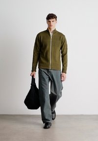 Olive green zip-up jacket with textured pattern, paired with loose-fitting gray pants; carrying a black fabric shoulder bag.