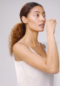Récourbe-cils en métal tenu à côté de l'œil, avec une personne portant un débardeur léger ; texture de peau douce, cheveux naturels, fond neutre.
