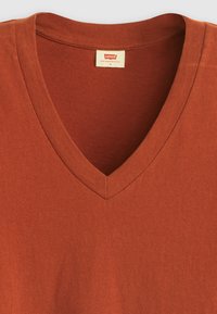 Tričko s výstřihem do V v rezavé barvě, vyrobené z měkké tkaniny s hladkým povrchem. Štítek uvádí "Levi's", velikost S. Jednoduchý, nezdobený design.