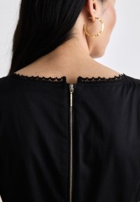 Zwarte jurk met een ronde halslijn, voorzien van kantafwerking, een onzichtbare ritssluiting aan de achterkant en een gladde stoftextuur. Grote gouden ringoorbel zichtbaar.