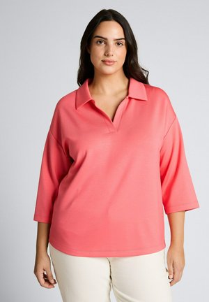 Kobieta z długimi ciemnymi włosami, ubrana w luźną koralowo-różową koszulę z kołnierzykiem oraz jasnobeżowe spodnie, stojąca na tle gładkiego szarego tła.