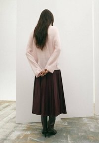 Suéter rosa claro con mangas anchas, combinado con una falda plisada burdeos oscura y botas negras de tacón hasta la rodilla. El cabello cae hacia atrás.