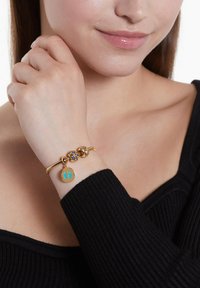 Goldfarbener Armband mit strukturiertem Band, dekorativen Perlen mit blauen Akzenten und einem runden Anhänger mit türkisfarbenen Fußabdrücken.
