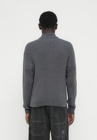 Άτομο που στέκεται με την πλάτη προς τον θεατή, φορά ένα ριμπ πλεκτό γκρι πουλόβερ και σκούρα cargo παντελόνια με τσέπες, πάνω σε απλό λευκό φόντο.