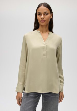 Donna che indossa una camicetta beige a maniche lunghe con scollo a V e polsini risvoltati, abbinata a jeans grigi in denim su uno sfondo semplice.