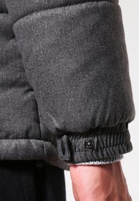 Mørkegrå dunjakkeærme med en ribbet manchet og en trykknap. Blød tekstur og quiltet design er tydeligt synligt.