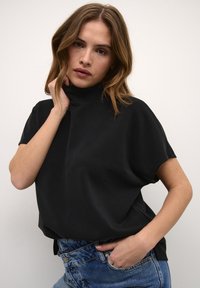 Schwarzes T-Shirt mit kurzem Ärmel und Stehkragen, aus weichem Material, mit lockerer Passform und glatterTextur, kombiniert mit hellblauen Jeans.