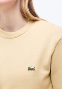 Γυναίκα που φοράει μπεζ φούτερ με μικρό πράσινο λογότυπο κροκοδείλου κεντημένο στην αριστερή πλευρά του στήθους.