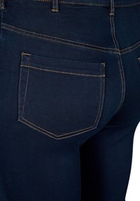 Los jeans de denim azul oscuro cuentan con un bolsillo trasero con costuras naranjas en contraste, una textura elegante y un diseño ajustado que enfatiza las líneas limpias.