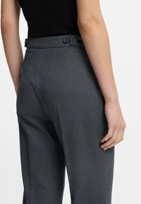 Primo piano di una persona che indossa pantaloni grigi scuri con texture, a vita alta e dettagli con bottoni, abbinati a un top nero.