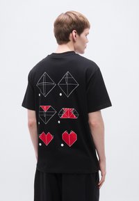 Μαύρο βαμβακερό μπλουζάκι με γεωμετρικά κόκκινα και λευκά σχέδια οριγκάμι στην πλάτη, με σχέδια διαμαντιού και καρδιάς, κοντά μανίκια και χαλαρή εφαρμογή.