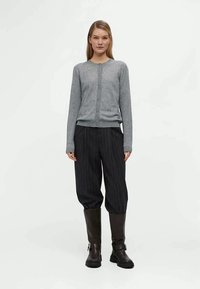 Γκρι πλεκτό κα cardigan με κούμπωμα, συνδυασμένο με μαύρο παντελόνι με ρίγες και καφέ δερμάτινες μπότες μέχρι το γόνατο. Το μοντέλο στέκεται μπροστά από έναν απλό φόντο.