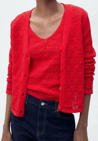 Roter Strickcardigan mit vorderer Knopfleiste und strukturiertem Design, getragen über einem passenden Tanktop. Kombiniert mit dunkelblauer Jeans.