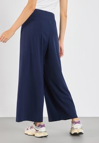 Pantalon large bleu marine en tissu texturé côtelé avec une ceinture élastique, associé à des baskets blanches et violettes.