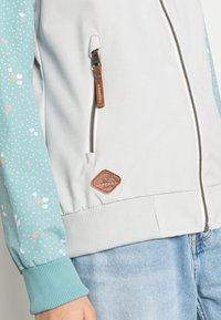 Casaco cinza claro com mangas em padrão floral teal, bolso com fecho éclair e punhos canelados. Apresenta um emblema de couro castanho na barra.