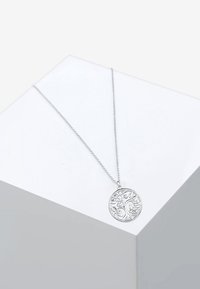 Sølvvedhængs halskæde med et cirkulært charms med detaljeret trædesign, hængende fra en delikat kæde på en hvid baggrund.