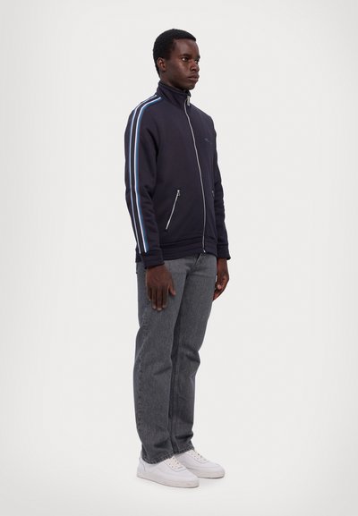 Svart zip-jacka med hög krage och blåa ränder, matchad med grå jeans och vita sneakers. Modellen står avslappnat mot en ljus bakgrund.