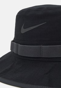 Gorra tipo bucket negra hecha de tela texturizada, con un logo de Nike en gris oscuro y una banda de tela a juego alrededor de la corona.