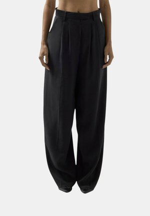 Persona che indossa pantaloni neri larghi a gamba larga con pieghe frontali, in piedi contro uno sfondo grigio chiaro uniforme.