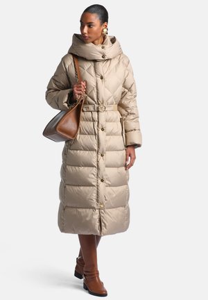 Femme portant un long manteau matelassé beige avec ceinture, portant un sac à main en cuir marron, et chaussant des bottines marron avec des boucles d'oreilles en or.