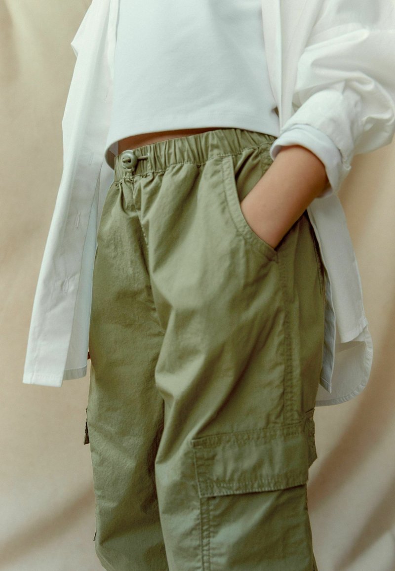 Personne portant un pantalon cargo ample vert olive, un haut court blanc et une chemise blanche déboutonnée, une main dans la poche, sur un fond neutre.
