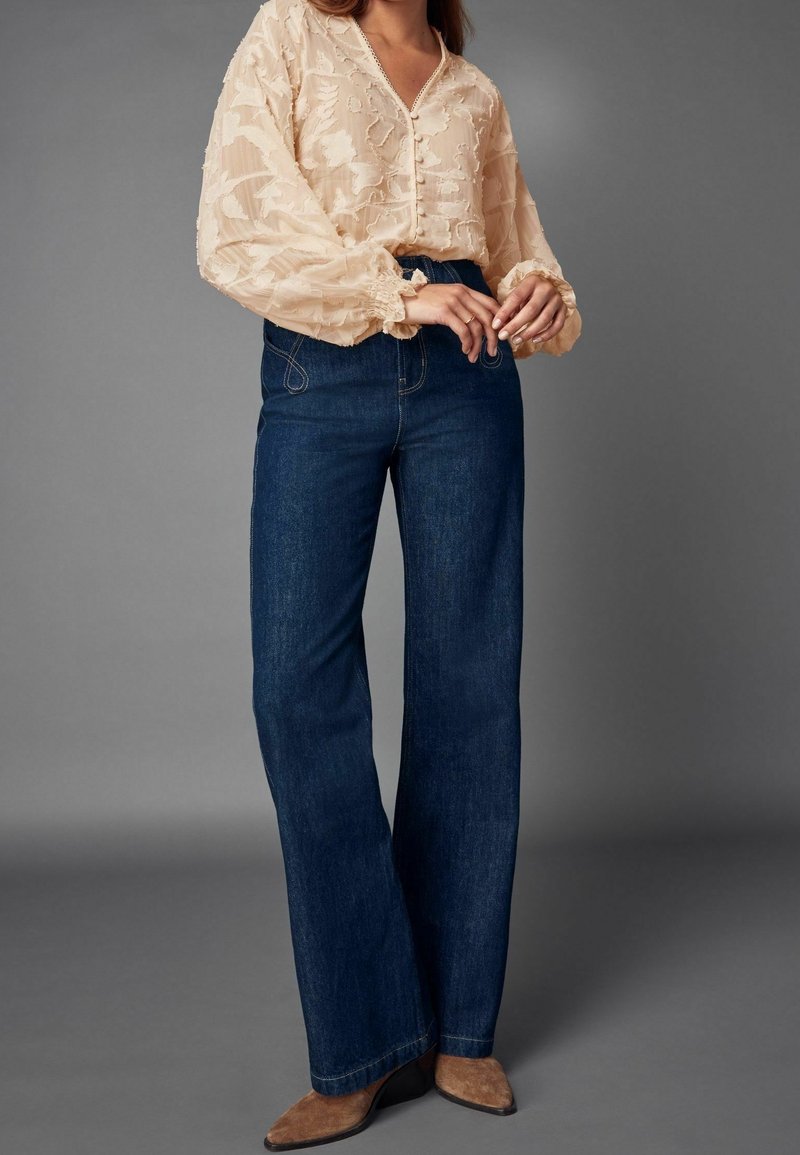 Femme portant un chemisier beige transparent à motifs floraux, un jean large taille haute bleu foncé et des chaussures pointues marron.
