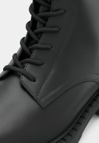 Bottine en cuir noir à lacets avec une texture lisse, un détail cousu en triangle et une semelle en caoutchouc plate avec un bord strié.