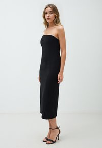 Schwarzes trägerloses Midikleid mit figurbetonter Silhouette, glatter Textur und einem Saum, der unter dem Knie endet. Kombiniert mit schwarzen High-Heels.