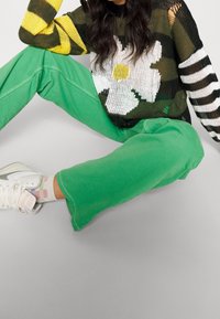 Pull tricoté avec motif floral, rayures vertes et jaunes ; associé à un pantalon vert. Baskets blanches et chaussettes à carreaux pastel visibles.