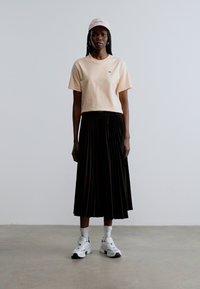 T-shirt courte rose clair, jupe midi plissée noire, baskets blanches et casquette rose clair. Fond uni, design minimaliste.