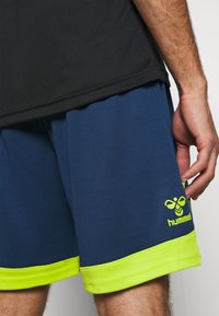 Hombre con pantalones cortos deportivos Hummel azul marino y amarillo neón y una camiseta negra, con la mano apoyada a su lado.