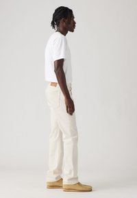 Hombre con el cabello trenzado que lleva una camisa blanca de manga corta, jeans color crema y zapatos de ante color miel, de pie de perfil sobre un fondo simple.