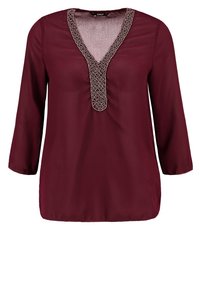 Blouse bordeaux avec un décolleté en V, des manches trois-quarts et un bord décoratif à motifs au niveau du décolleté. Texture de tissu douce et légère.