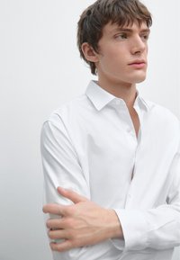 White long-sleeved button-up shirt with a classic collar, smooth texture, and fitted design. Worn with an open collar, minimalistic look.