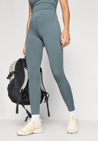 Legíny v tlmenej teal farbe, vyrobené z elastického materiálu. Vhodne doplnené čiernym ruksakom a béžovými teniskami s bielymi ponožkami.