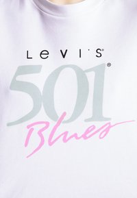 Tissu blanc avec le logo "Levi's" en lettres noires, un grand "501" gris et le mot "Blues" en pink écriture cursive imprimés sur le devant.
