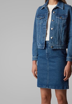 Femme portant une veste en jean bleu avec des boutons en métal et une jupe en jean bleu assortie, debout devant un fond gris uni.