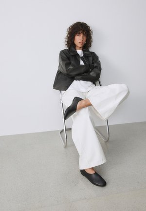 Persona con cabello rizado, que lleva una chaqueta de cuero negra, pantalones blancos y zapatos negros, sentada con las piernas cruzadas en una silla metálica plateada contra una pared lisa.