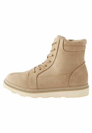 Beige mockalädersankläder med snörning, rund tå, vadderad krage och en vit gummisula med en sömdekoration längs kanten.