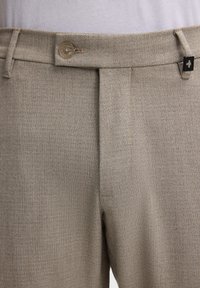 Beige strukturierte Hose mit flacher Vorderseite, einreihigem Knopfverschluss und einem kleinen dekorativen schwarzen Akzent am Bund.