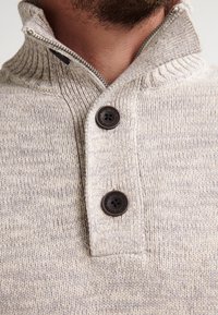 Persona con barba che indossa un maglione lavorato a maglia beige con colletto con zip e due bottoni marrone scuro sulla parte anteriore.