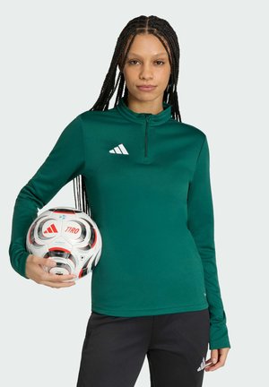 Kobieta z długimi warkoczami, ubrana w zielony top Adidas i czarne spodnie, trzymająca biało-czerwoną piłkę nożną Adidas na wysokości talii.