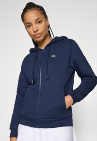 Marine zip-up hettegenser i myk stoff med snor, med en liten grønn logo på brystet og frontlommer. Glatt tekstur.