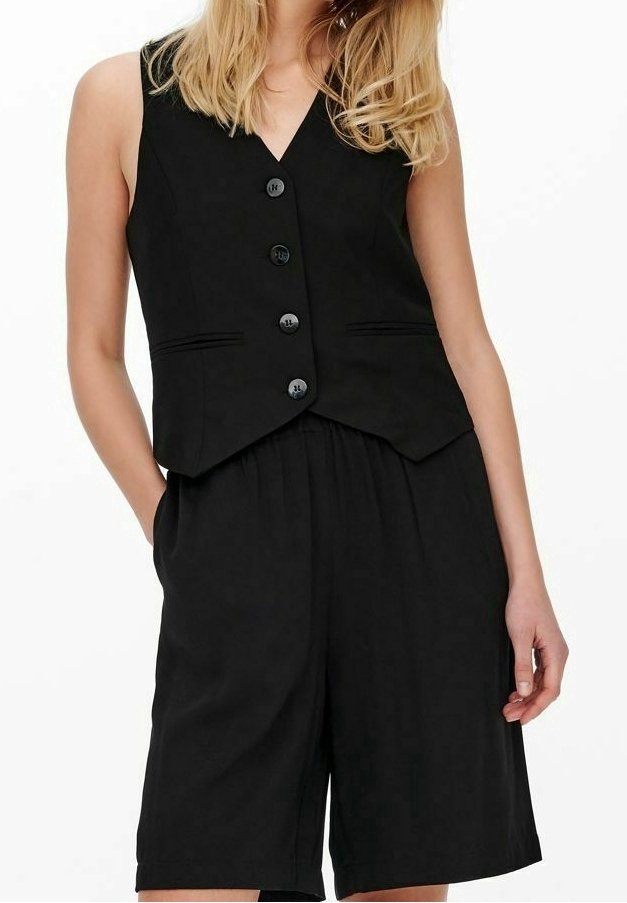 Femme portant un gilet noir sans manches boutonné et un short ample taille haute assorti, les mains dans les poches, sur fond blanc.