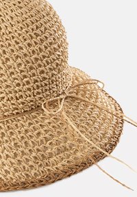 Kapelusz słomkowy z szerokim rondem, o teksturowanym, plecionym wzorze, w jasnym odcieniu beżu, z przewiązaną dekoracją ze słomy u podstawy.