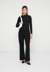 Tânără îmbrăcată într-un pulover negru cu dungi și pantaloni largi, ținând o geantă albă de umăr, stând în fața unui fundal alb simplu.