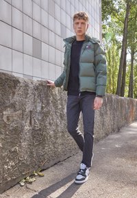 Chaqueta verde, esponjosa, con capucha y logo, camiseta negra, jeans oscuros, zapatillas en blanco y negro, de pie contra una pared texturizada a la luz natural.