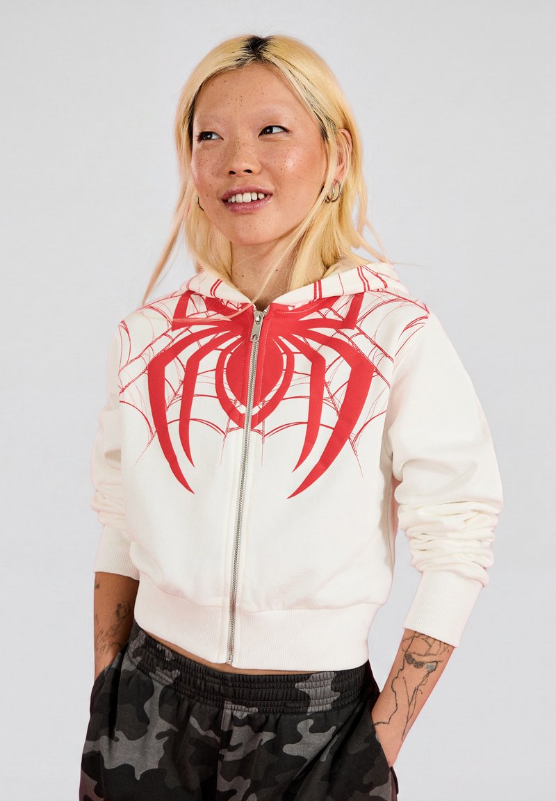 Young woman with blonde hair wearing white zip-up hoodie with large red spider design and black camo pants, hands in pockets, looking right.