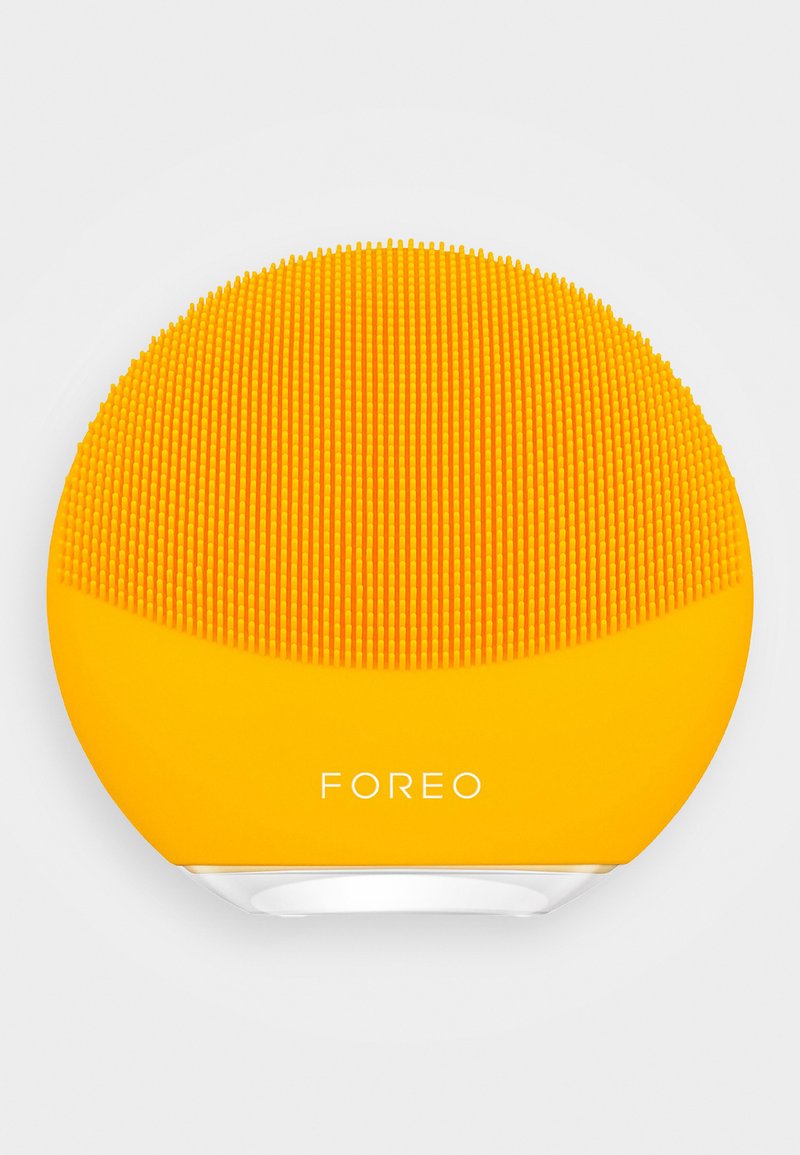 Silikon ansiktsrensingsapparat, rund form, lys gul, strukturert med små riller, har merkevarenavnet "FOREO" på fronten.